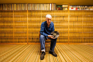 Elderly man holding vinyl record in front of large vintage record collection shelves