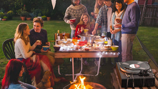 Family enjoying outdoor dinner with record player, fire pit, and festive lights in backyard