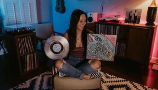 Woman holding vintage clear vinyl record and colorful album cover in a cozy music room with turntables and records.